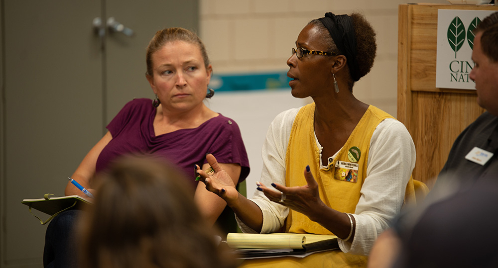 A Black woman in a yellow dress gestures with her hands and speaks. To her left, a White woman in maroon listens.