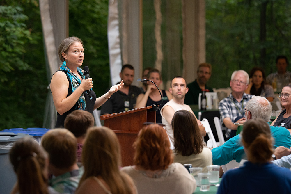 A White woman stands at a podium, holding a microphone and gesturing with her hand. In the foreground and background, a seated crowd faces the speaker.