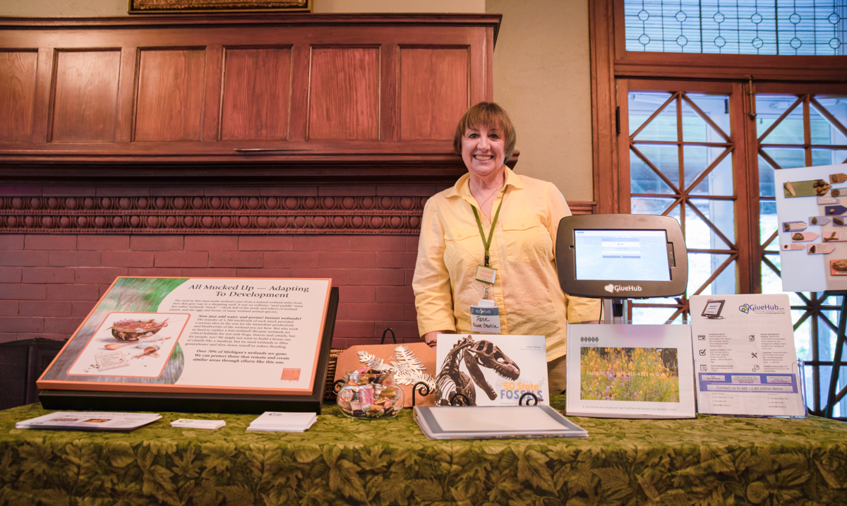 A table with an assortment of cards, brochures, booklets, and signs. Behind the table stands a smiling White woman in a yellow shirt. In the background are painted bricks, wooden cabinets, and a glass door.