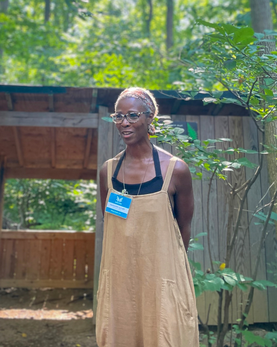 A Black woman in a beige dress and glasses stands outside, illuminated by sunshine from behind. In the background is a wooden structure and a blurry leafy woodland scene.