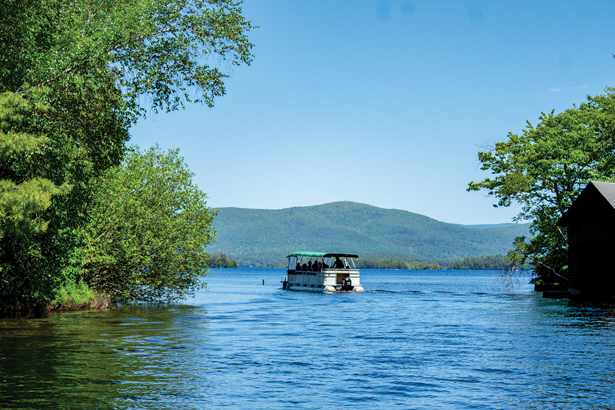 A pontoon boat in water. To the left and right are shrubs and trees in green foliage. In the distance, forested hills with blue sky above.