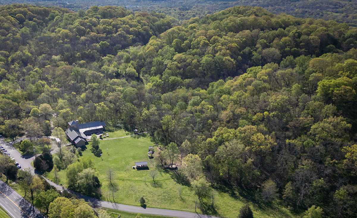 An aerial view of a hilly forested landscape. In the bottom-left corner, a building stands beside a field of open grass and a curved road.