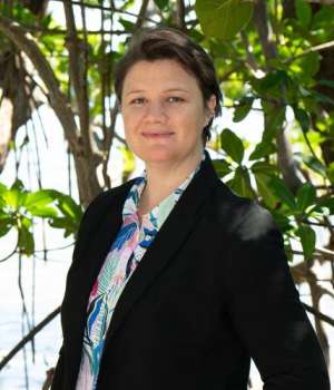 A person stands outside with green foliage behind. They wear a black blazer over a white shirt with a multi-colored floral motif. A person stands outside with green foliage behind. They wear a black blazer over a white shirt with a multi-colored floral motif.