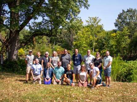 Two rows of people pose in a lawn outside. Behind them are trees and shrubs that surround a pond. Two rows of people pose in a lawn outside. Behind them are trees and shrubs that surround a pond.
