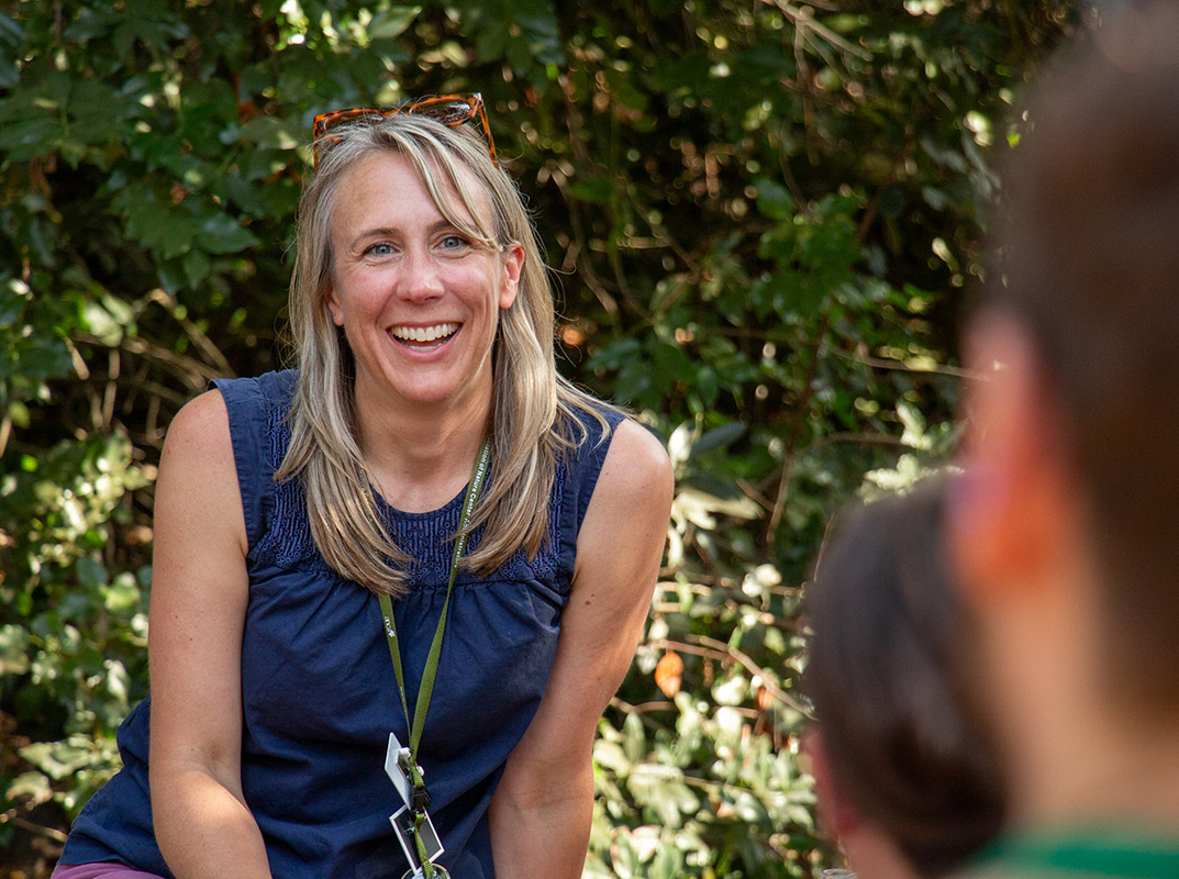 A person smiles at people in the foreground. Behind them, green foliage.